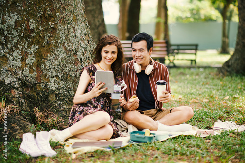 Cheerful young diverse couple watching funny videos on tablet computer when having picnic in park