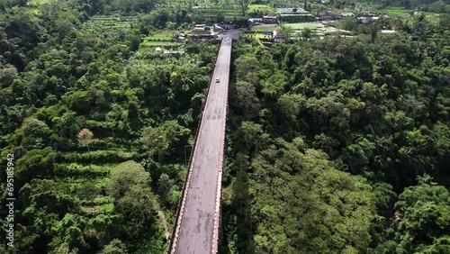 Scenic aerial shot of Jembatan Tukad Bangkung, large balanced cantilever bridge crossing tropical ravine. Orbiting motion of flying camera. Only few vehicles ride by at empty roadway photo