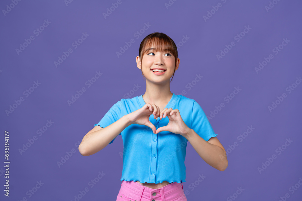 Young asian woman shows her fingers heart-shaped sign and presses to chest on purple background. Young asian woman shows her fingers heart-shaped sign and presses to chest on purple background.