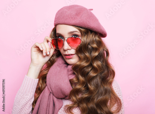 Young woman with curly hair wearing a pink sweater, scarf, and beret over pink background. © Raisa Kanareva