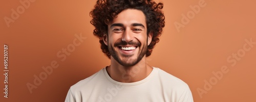 A handsome and cheerful man with curly hair, wearing a toothy smile, posing happily against a brown background in a portrait. © Andrey