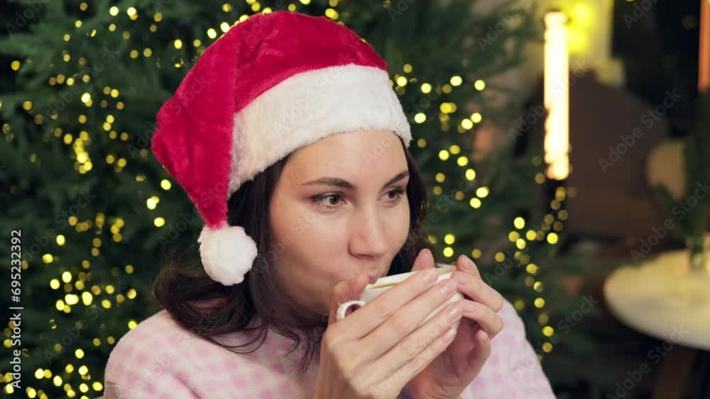 An attractive young woman in a red Christmas hat and pajamas takes a sip of hot coffee and looks in front of her An attractive young woman in a red Christmas hat and pajamas takes a sip of hot coffee and looks in front of her