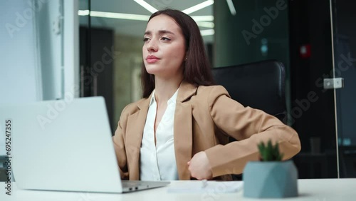 Front view of overtired caucasian businesswoman working with wireless laptop in own cabinet. Exhausted female dressed in formal suit feeling severe back pain while sitting on office chair indoors. photo