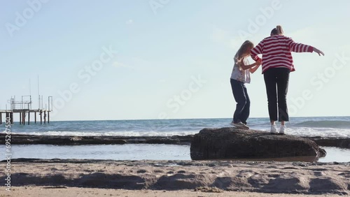 Mom and daughter by the sea happily running towards big stone on seashore. Young woman in striped T-shirt with her little girl jumping on a rock in a sunny day, family active day by the seaside