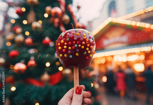 close-up photo of hand holding a caramel apple sprinkled with candies, glazed apple on a wooden stick, Christmas tree and Christmas market on background 
