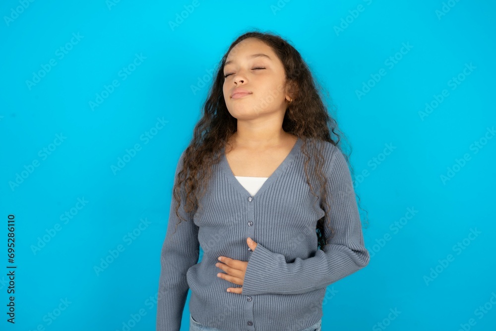 Beautiful teen girl wearing blue jacket over blue background touches tummy, smiles gently, eating and satisfaction concept. Beautiful teen girl wearing blue jacket over blue background touches tummy, smiles gently, eating and satisfaction concept.