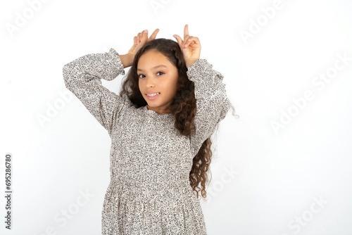 Funny beautiful caucasian teen girl wearing flowered dress shows horns, fingers on head gesture, posing silly and cute