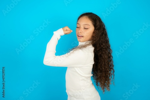 Portrait of funny Young beautiful teen girl wearing white turtleneck over blue background shout yeah raise fists hands celebrate victory game competition