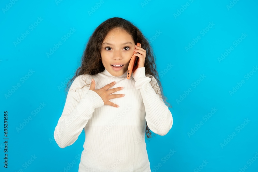 Smiling Young beautiful teen girl wearing white turtleneck over blue background talks via cellphone, enjoys pleasant great conversation. People, technology, communication concept Smiling Young beautiful teen girl wearing white turtleneck over blue background talks via cellphone, enjoys pleasant great conversation. People, technology, communication concept