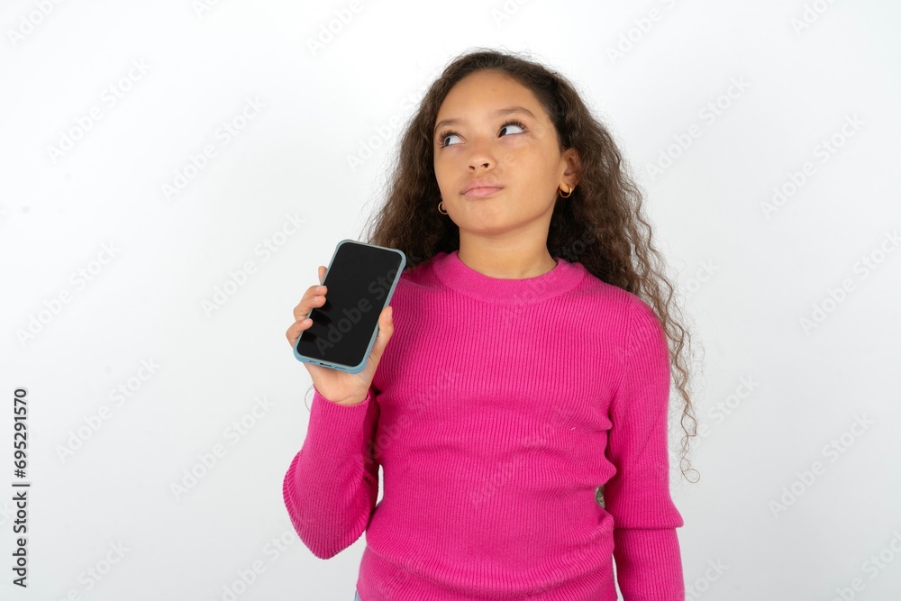 Beautiful teen girl wearing pink sweater over white background holds new mobile phone and looks mysterious aside shows blank display of modern cellular Beautiful teen girl wearing pink sweater over white background holds new mobile phone and looks mysterious aside shows blank display of modern cellular