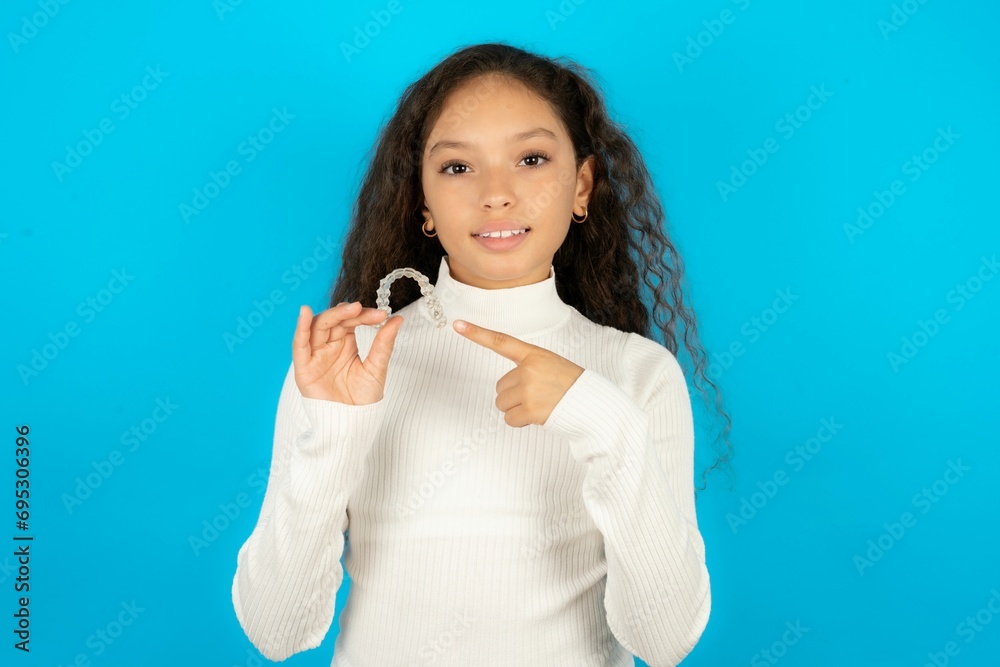 Beautiful kid girl standing over blue background holding an invisible aligner and pointing at it. Dental healthcare and confidence concept. Beautiful kid girl standing over blue background holding an invisible aligner and pointing at it. Dental healthcare and confidence concept.