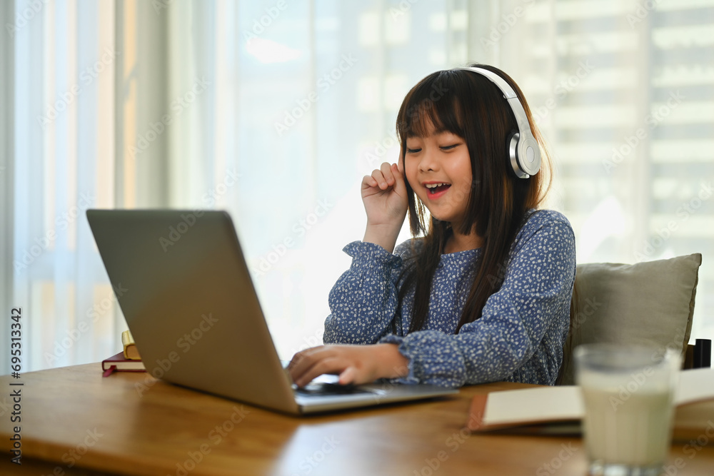 Smiling Asian little cute girl in headphone watching online lesson on laptop at home. Smiling Asian little cute girl in headphone watching online lesson on laptop at home.