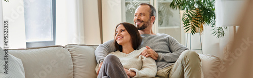 cheerful child-free couple sitting on cozy sofa in living room and looking away, horizontal banner photo