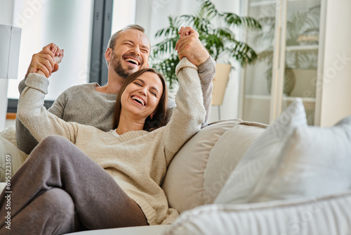 cheerful child-free couple holding hands and laughing with closed eyes on couch in living room, fun photo