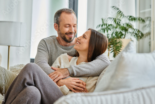 joyful child-free couple embracing and looking at each other on couch in living room, love and joy photo