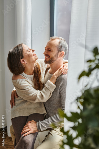 laughing child-free couple embracing and smiling at each other near window at home, tenderness