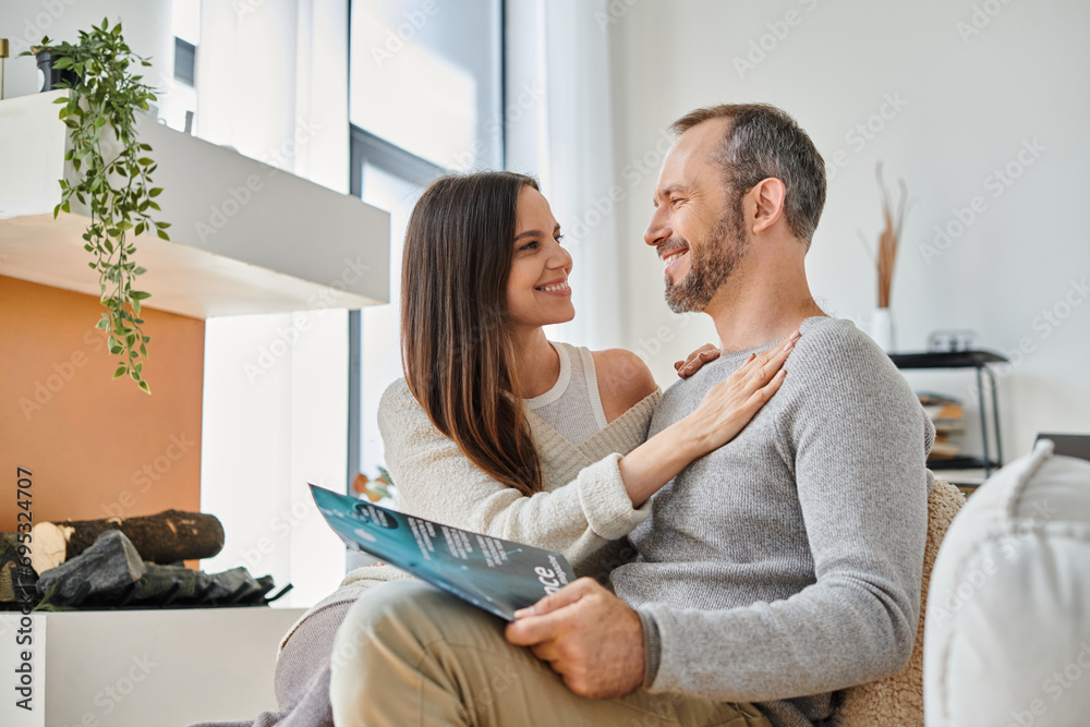 smiling woman embracing husband reading science magazine on couch at home, child-free life smiling woman embracing husband reading science magazine on couch at home, child-free life