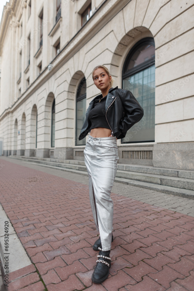 Beautiful street fashion girl model in stylish clothes with a leather jacket and skirt stands in the city near a vintage building