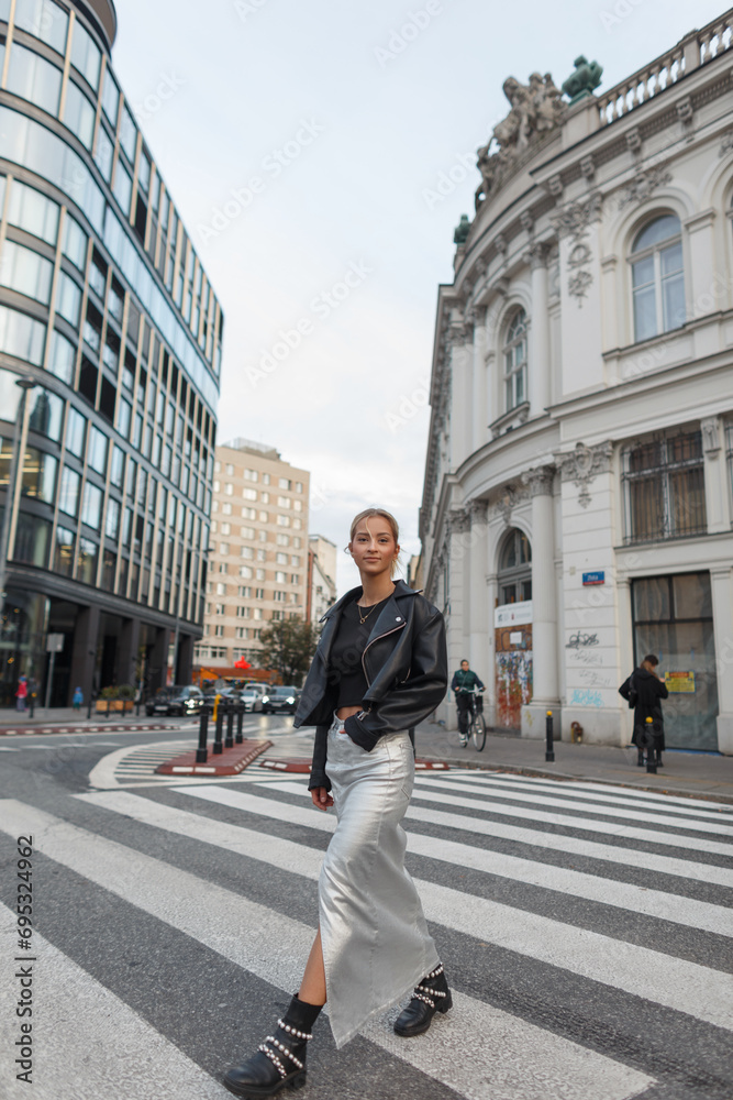 Fashion beautiful girl in stylish urban clothes with a leather jacket and skirt walks through a crosswalk on the street near buildings