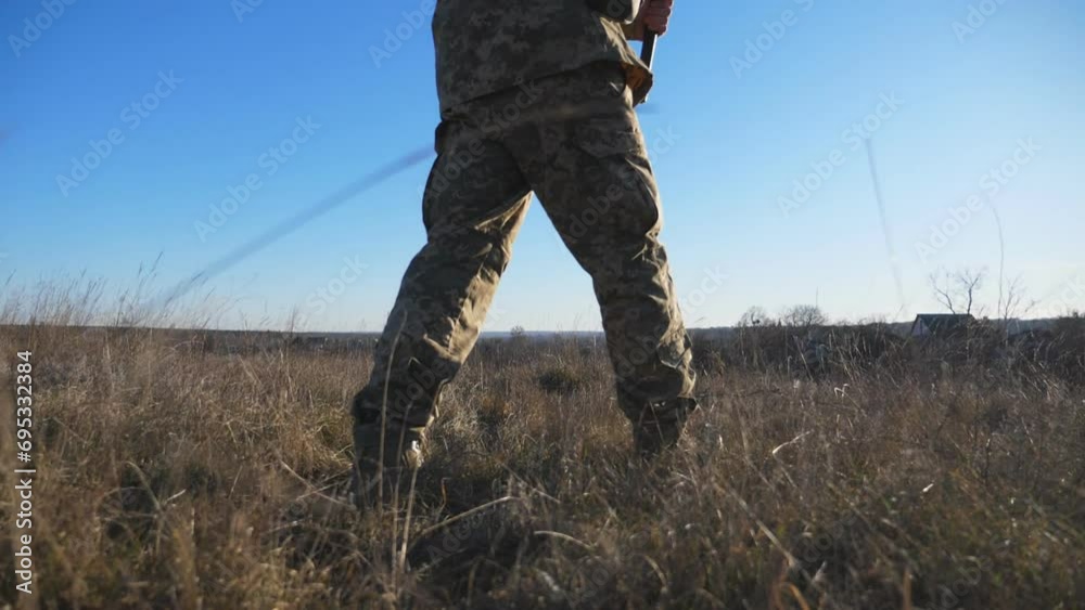 Dolly shot of legs of young army man in military shoes walking along field on sunny day. Male feet of ukrainian soldiers going in brown boots through dry grass at countryside. War in Ukraine. Slow mo