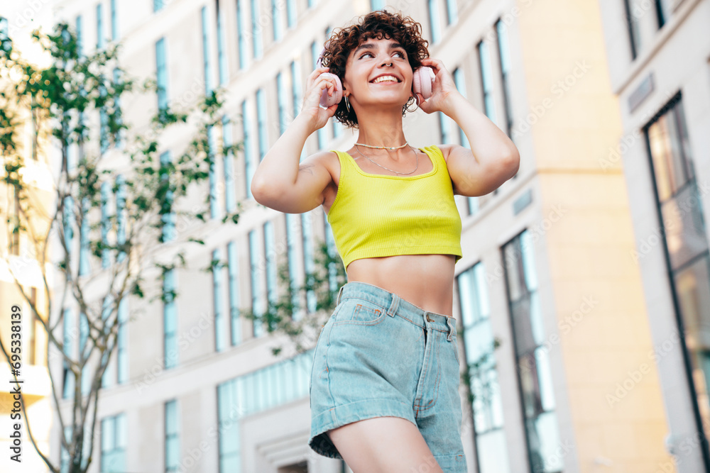 Young beautiful smiling hipster woman in trendy summer clothes. Carefree woman with curls hairstyle, posing in the street at sunny day. Positive model outdoors. Listens music at her headphones