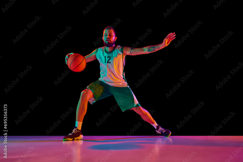 Full-length portrait of basketball player honing skills with precision dribbling and making powerful slam dunk against black background in neon.