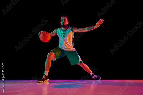 Full-length portrait of basketball player honing skills with precision dribbling and making powerful slam dunk against black background in neon.