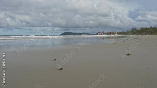 The waves of the ocean roll towards the shore. On the beach, there are bunches of algae, tiny crabs running around. In the distance, among the green vegetation, the hotel is visible. Clouds, blue sky