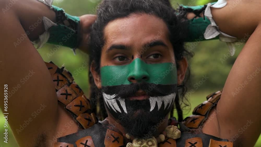Bearded male warrior with painted face fixing dreadlocks wearing neck costumes while standing in green park with trees