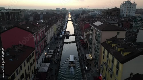 From above drone in motion of the Navigli district in Milan at sunset, with a canal boat cruising down the waterway and people walking on the vibrant canal-side walkways photo