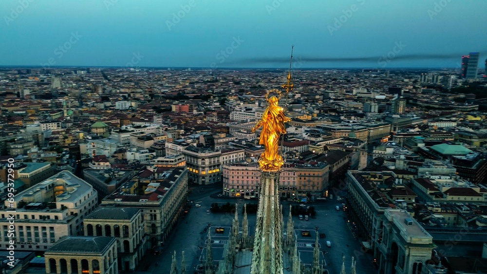 Aerial view of the statue of the Madonna on the central spire of the Duomo and the city's cathedral at dawn. Roofs of houses and skyscrapers. Sun over the horizon. duomo square. Italy Milan 16.11.2023 - obrazy, fototapety, plakaty 