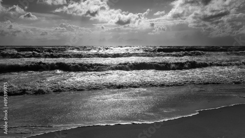 Panorama with waves on a stormy sunny summer day on the beach of Sylt island, Germany. Cloudy sky in holiday spot Westerland with spray, foam and reflections on North Sea. Black and white greyscale.