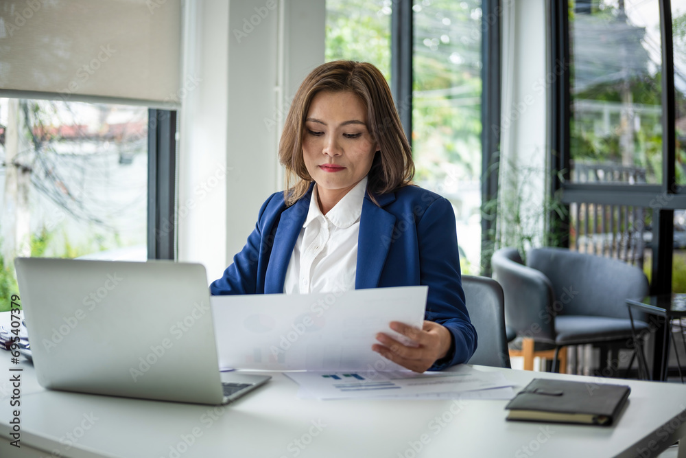 Businesswoman sitting at desk on couch in workplace or at home working on laptop and analyzing data on charts and graphs and writing on papers Businesswoman sitting at desk on couch in workplace or at home working on laptop and analyzing data on charts and graphs and writing on papers