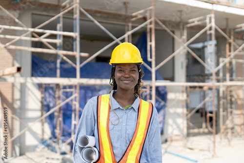 Portrait of African American female civil engineer wearing safety vest with helmet standing and hold blueprint building plan at construction site outside