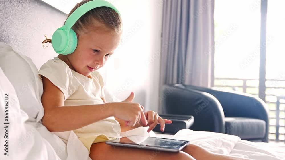 Little girl in headphones using digital tablet while listening to music or playing game in home bed Little girl in headphones using digital tablet while listening to music or playing game in home bed