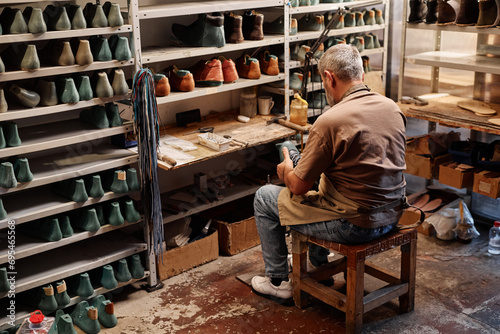 Back view of mature man making pair of new shoes or boots in workshop while sitting on stool in front of shelves with workpieces