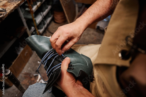 Hands of professional male shoemaker putting shoelace into upper part of unfinished black leather boot while sitting in workshop photo