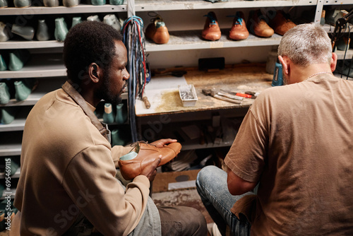 Young African American man in workwear looking at shoemaking master while sitting next to him in workshop with workpieces on shelves photo