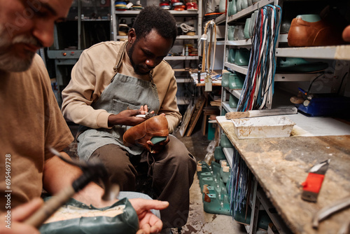 Young African American man in apron putting together upper part of footwear with sole while working over new pair of shoes or boots