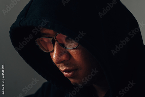 A male hacker wearing glasses and a hoodie covers his head in the dark is hacking into a victim's financial system. Data thief, internet fraud, darknet and cyber security concept.