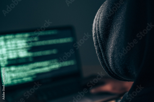 Behind the scenes of an anonymous hacker wearing a hoodie over his head in the dark hacking into a victim's financial system. Data thief, internet fraud, darknet and cyber security concept.
