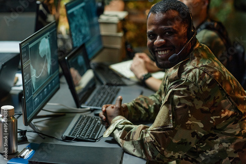 Happy young African American male military officer with headset looking at camera with smile while sitting by workplace with computers