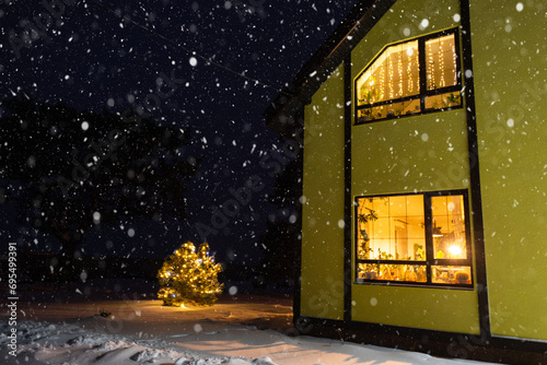 Cozy festive window of the house outside with the warm light of fairy lights garlands inside - celebrate Christmas and New Year in a warm home. Christmas tree, bokeh, snow on pine trees and snowfall