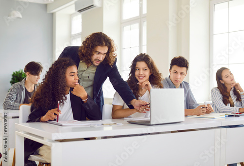 Group of high school students and classmates sitting in the classroom with their young man teacher and looking at the laptop monitor screen during a lesson. Education and back to school concept.
