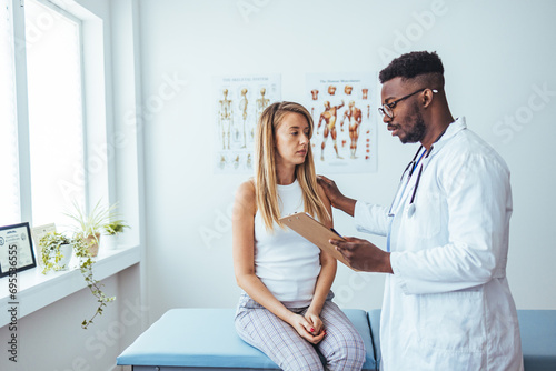 Doctor and patient talking during medical checkup at clinic. Young man in white coat showing and explaining analysis results to woman sitting on medical couch in examination room photo