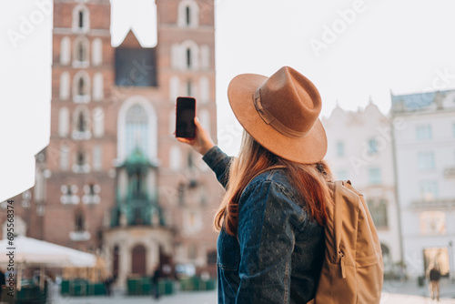 Happy young European tourist with backpack in hat makes photo or video on smartphone on Market Square in Krakow. Traveling Europe in summer. St. Marys Basilica. Vacation concept photo