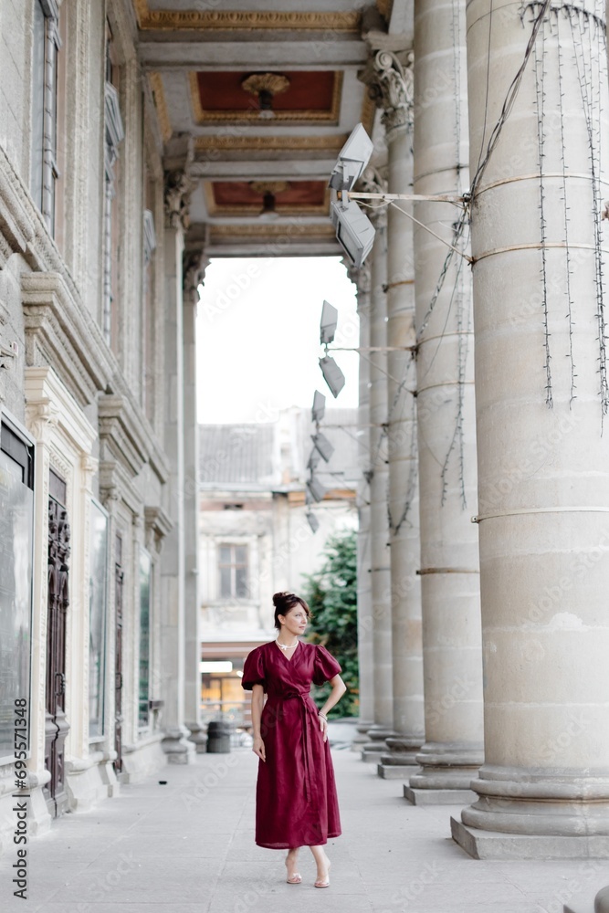 Fototapeta premium Attractive girl with hairstyle, in red dress is walking on street on old building background