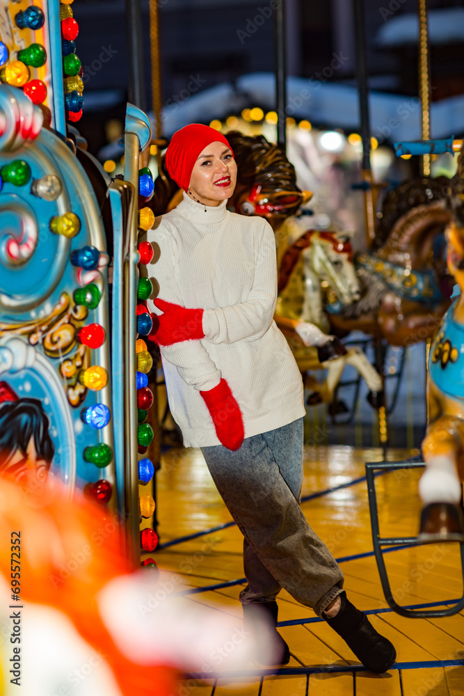 A young, beautiful woman walks in the park in an amusement park, rides on carousels, wearing a hat and winter clothes.