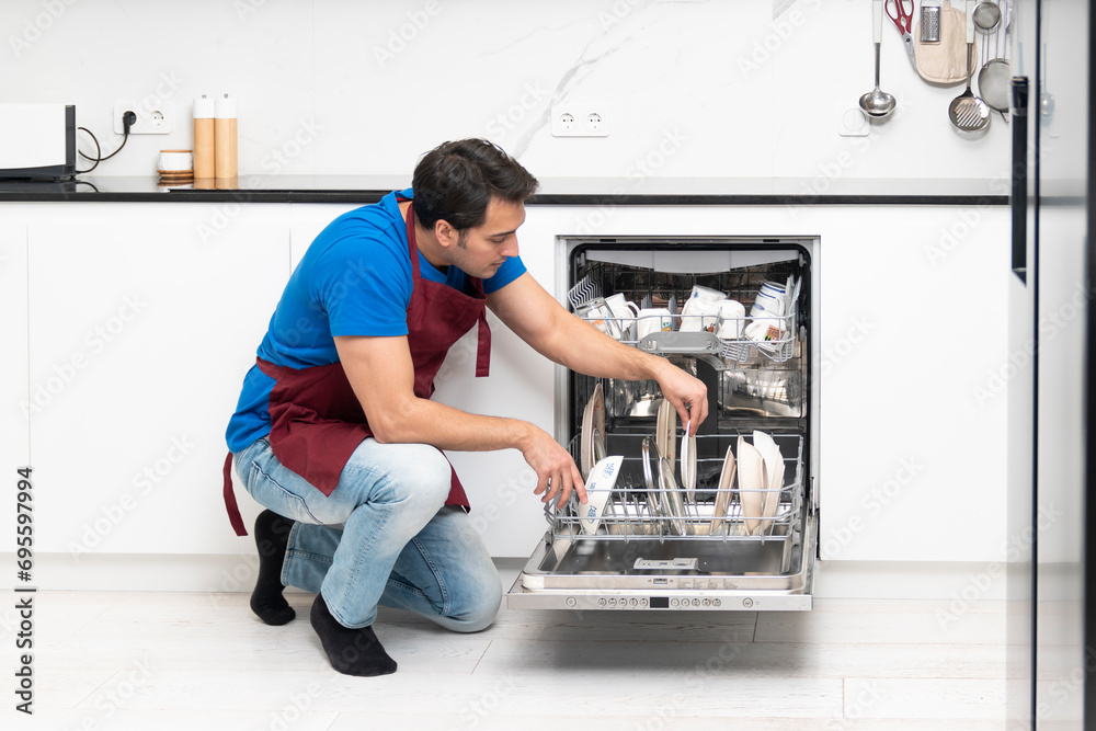 Young handsome man putting dirty plates in dishwasher machine in the kitchen. Household and exhausting cleaning day concept Young handsome man putting dirty plates in dishwasher machine in the kitchen. Household and exhausting cleaning day concept