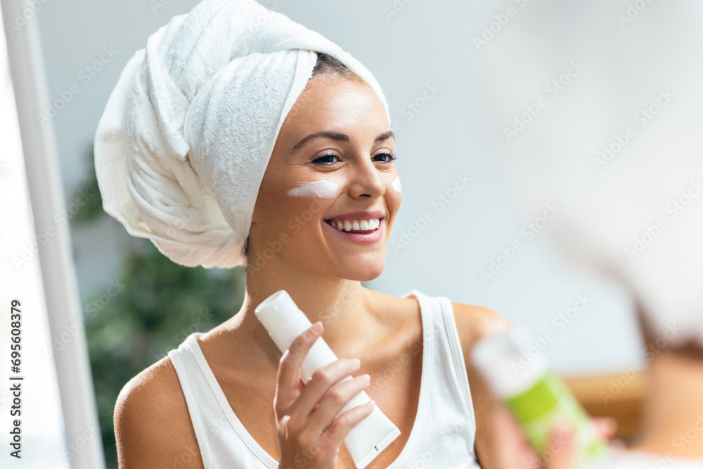 Beautiful young woman caring of her skin while putting on cream looking at mirror at home. Beautiful young woman caring of her skin while putting on cream looking at mirror at home.
